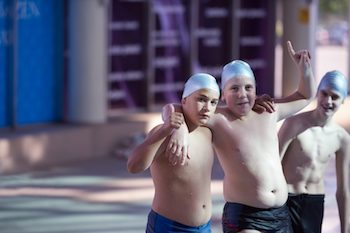 Three boys standing near an indoor pool wearing swim caps